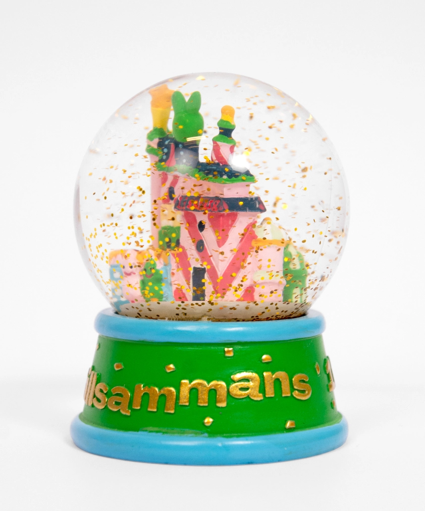 En närbild av en Liseberg-snöglob som visar ett miniatyrhus i rosa och grönt, med guldglitter som svävar i globen. Den gröna basen har en blå kant och ordet "tillsammans" i guldtext.